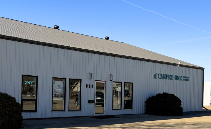 A white metal building with a gray roof, featuring multiple windows and the "CARPET ONE FLOORING" logo. A dark green bush is planted in front of the building.